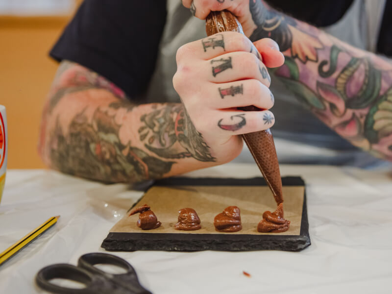 A close up of hands with tattoos piping chocolate truffles onto a baking tray